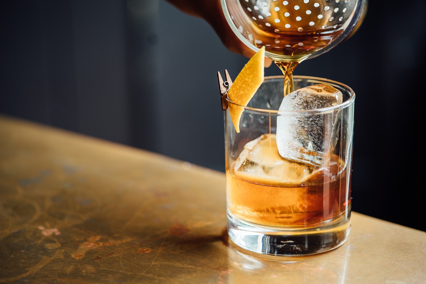 Ein Aperitif wird über einen Eiswürfel gegossen, daneben eine Orangenzeste